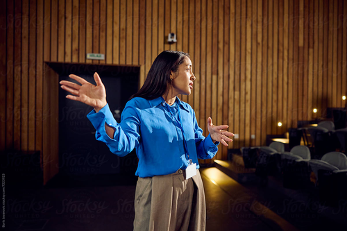 A woman in a blue blouse and tan pants gestures expressively while speaking in a wood-paneled auditorium. She wears a name badge and appears to be delivering a presentation or speech on stage.