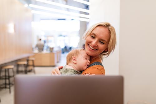 A smiling blonde woman holds a sleeping baby against her chest while looking at a laptop in a bright, modern café setting.