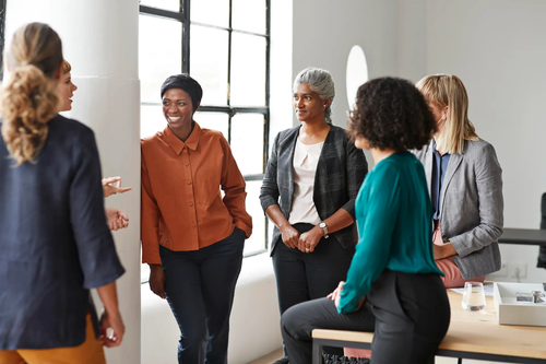 A diverse group of five professional women engaged in conversation in a modern office setting, dressed in business casual attire, appearing relaxed and engaged in a friendly workplace discussion.