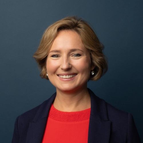 A professional headshot of a smiling woman with short blonde hair, wearing a red top and navy blazer, against a dark blue background. She has pearl earrings and a warm, confident expression.