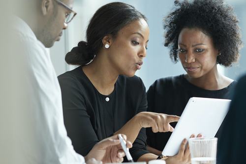 Three colleagues collaborate around a tablet, with a woman in black pointing at the screen while explaining something to a man in white and another woman with natural hair looking on attentively.