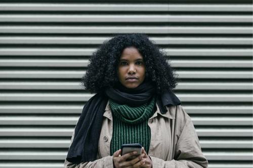 A young Black woman with curly hair stands against a corrugated metal wall, wearing a beige coat, black scarf, and green sweater. She holds a smartphone and gazes seriously at the camera.