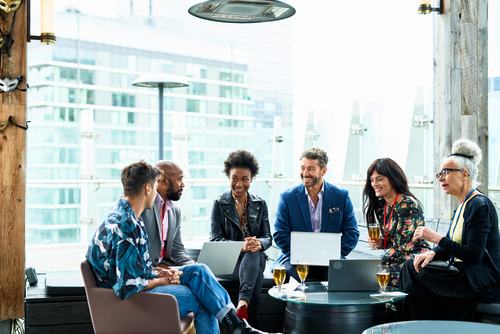 A diverse group of six professionals networking at a rooftop venue, gathered around a table with laptops and champagne glasses, engaged in lively conversation against a city skyline backdrop.