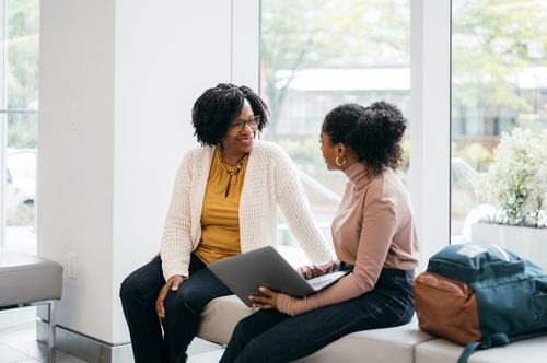 Two women sit together in a bright, modern space, engaged in conversation. One smiles warmly while the other holds a laptop. Bags rest nearby, suggesting a casual meeting or mentoring session in an office or campus setting.