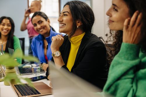 A group of diverse women smiling and engaged in a meeting or workshop, seated around a table with laptops. A woman in a yellow turtleneck and black blazer is prominently featured, looking attentive and cheerful.