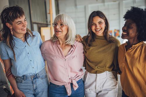 Four diverse women of different ages smile and laugh together, arms around each other, in a bright indoor setting. They wear casual, colorful tops and share a joyful, close moment.