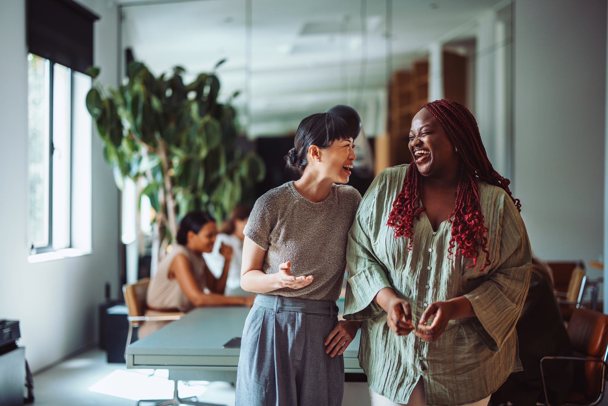 Two women laugh together in a bright, modern office. One wears a gray top and trousers; the other wears a green blouse with red braids. A colleague works in the background near large windows and lush plants.
