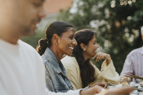 A group of people smiling and laughing during an outdoor gathering, seated at a table with food. A woman in a denim jacket is prominently featured, enjoying the moment with friends or family amid lush greenery.