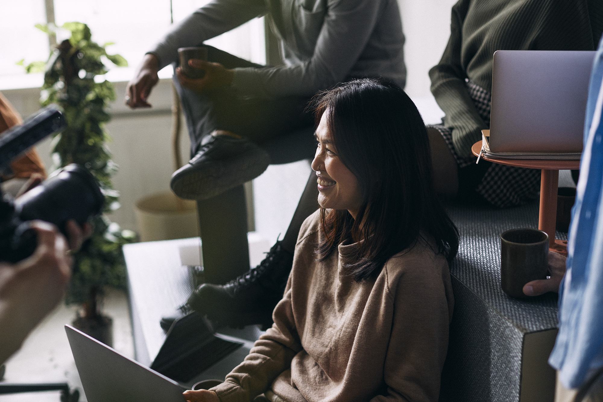 A smiling woman in a beige sweater sits with a laptop during a casual office gathering, surrounded by colleagues with coffee cups and laptops, while someone films with a camera nearby.