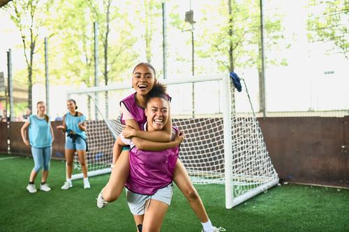 Two girls in purple vests celebrate joyfully on an outdoor soccer field, one giving the other a piggyback ride near the goal. Teammates cheer in the background on a sunny day.