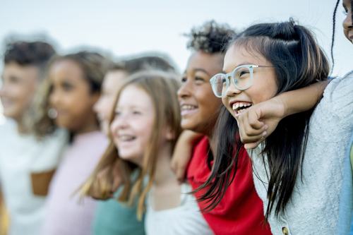 A diverse group of happy children laughing and smiling together outdoors. A girl wearing glasses is in focus in the foreground, with other kids standing beside her in a joyful, candid moment.