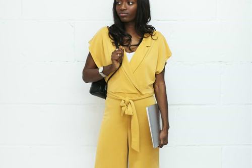 A stylish young woman wearing a mustard yellow wrap jumpsuit with wide-leg pants, holding a laptop, carrying a black bag, and wearing a watch, posed against a white brick wall.