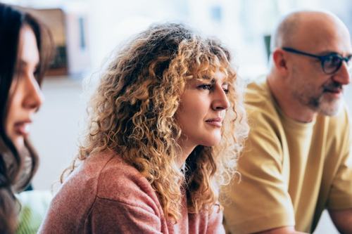 Three people sit together indoors. A woman with curly blonde hair in a pink top is in focus at center, looking ahead thoughtfully. A dark-haired woman is at left, and a bald man with glasses in a yellow shirt sits at right.
