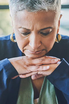 An older woman with short gray hair and gold hoop earrings looks downward with a thoughtful, somber expression, her hands clasped together beneath her chin, wearing a blue jacket.
