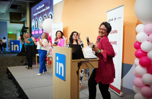 A lively Lean In Latinas panel event at LinkedIn, featuring women on stage celebrating and clapping near a podium with the LinkedIn logo, surrounded by pink and white balloons.