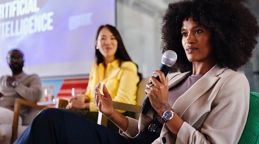 A woman with an afro speaks into a microphone at an Artificial Intelligence panel discussion, while colleagues listen attentively behind her, with a purple AI-themed presentation screen visible in the background.