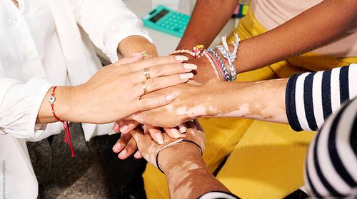 A diverse group of people stacking their hands together in a unity gesture, wearing colorful bracelets and rings, symbolizing teamwork and togetherness.