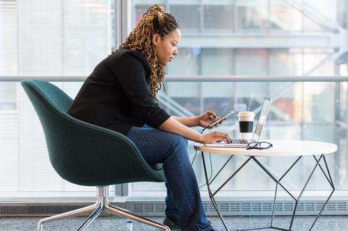 A woman with locs sits in a teal chair by large windows, working on a laptop while holding a smartphone. A coffee cup and glasses rest on a modern geometric table beside her.