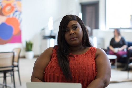 A woman in a red sleeveless top sits with a laptop in a bright, modern workspace, looking thoughtfully upward. A colorful abstract artwork hangs on the wall behind her, with another person seated in the background.